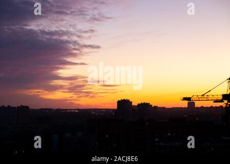 Picturesque landscape in the city at dawn with multi-colored yellow and purple sky and silhouettes of houses and a high crane at a construction site. Stock Photo