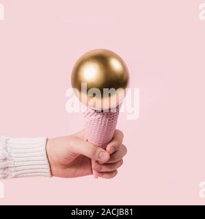 hand in white sweater holds pink ice cream cone with golden ball. Minimal Christmas concept. Stock Photo