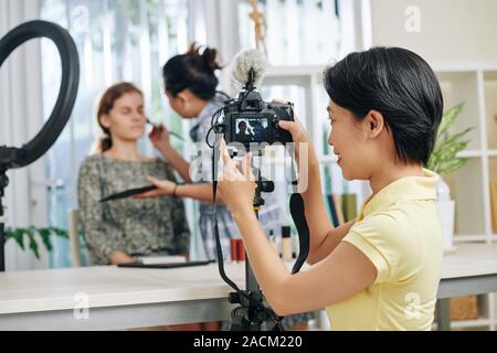 Smiling young Asan woman setting camera to shoot video of make-up artist working with model Stock Photo