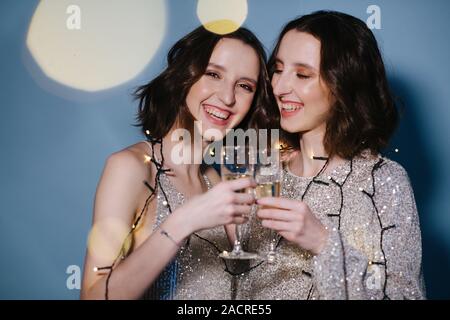 Twin sisters in shiny evening dresses celebrate with glasses of sparkling wine. Stock Photo