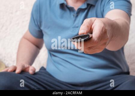 White middle-aged man in a blue t-shirt with a remote control from the TV. A man on a cozy sofa switches television channels Stock Photo