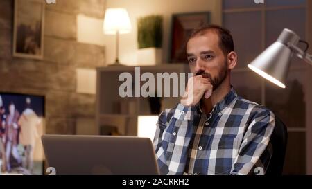 Businessman thinking and holding arms crossed while working on laptop in home office during night hours. Stock Photo