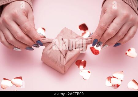 Gift in female hands with a beautiful manicure. Gift in ecological packaging - Furoshiki. Festive background with hearts. Stock Photo