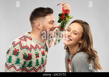 christmas, people and holiday traditions concept - portrait of happy couple in ugly sweaters kissing under the mistletoe over grey background Stock Photo