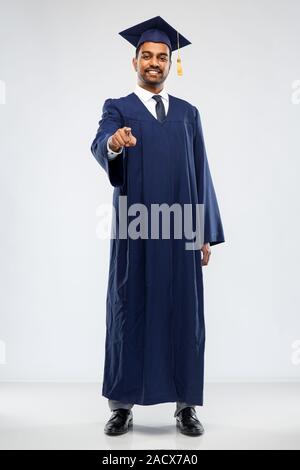 education, graduation and choice concept - happy smiling indian male graduate student in mortar board and bachelor gown pointing to you over grey back Stock Photo