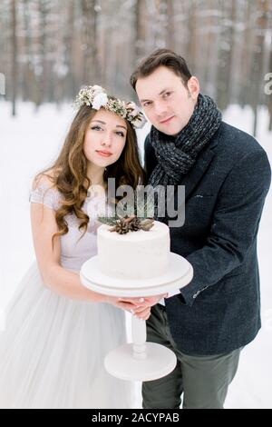 bride in flower wreath and stylish groom holding winter wedding decorated cake. Winter wedding in snowy forest Stock Photo