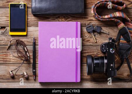 Overhead view of travel gear placed on wooden table. Mobile phone, earplugs, violet sketchbook, pencil, camera and purse. Flat l Stock Photo
