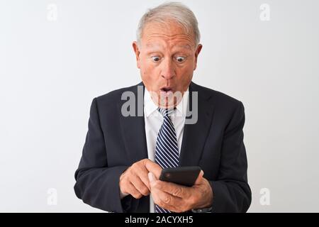 Senior grey-haired businessman using smartphone standing over isolated white background scared in shock with a surprise face, afraid and excited with Stock Photo