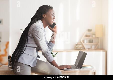 Young Black Businesswoman Talking On Cellphone And Using Laptop In Office Stock Photo