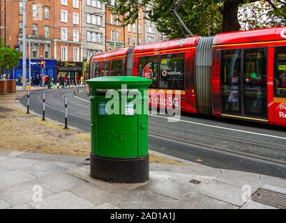 Green mail post box for Irish post service provider An Post, in Dublin city centre, with LUAS tram light rail system passing by. Transport Ireland Stock Photo