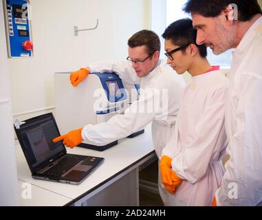 Genetic PCR analysis. Researchers examining results on a laptop (lower left) connected to a real-time polymerase chain reaction (PCR) machine (centre Stock Photo