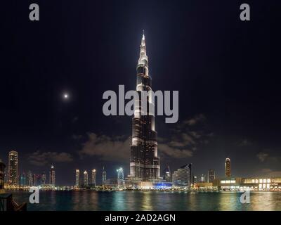Burj Khalifa skyscraper, Dubai, United Arab Emirates, at night with surrounding buildings. The Moon at left in the immensely light-polluted sky. Const Stock Photo