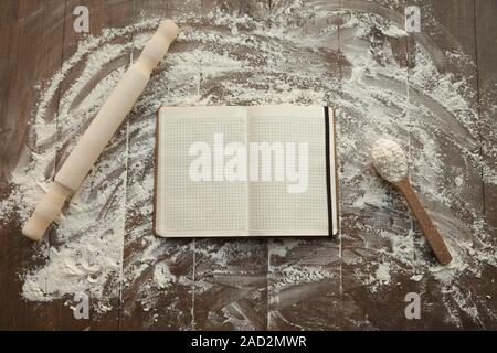 Composition of clear open recipe book in middle and rolling pin with spoon by sides on floured table. Stock Photo