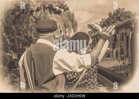 Black white rear back view of UK couple, 1940s dress, on platform, Severn Valley Railway ww2 station waving goodbye to friends on vintage steam train. Stock Photo