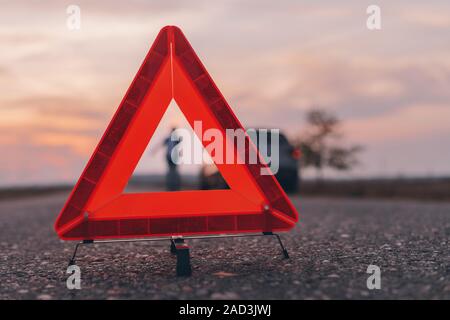 Warning triangle sign on the road, woman in blur background calling for roadside assistance by the broken car, selective focus Stock Photo