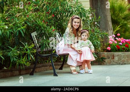 Beautiful young mom sits with her daughter in the park. Pink dresses. They are happy and laughing. Nice warm weather. Stock Photo