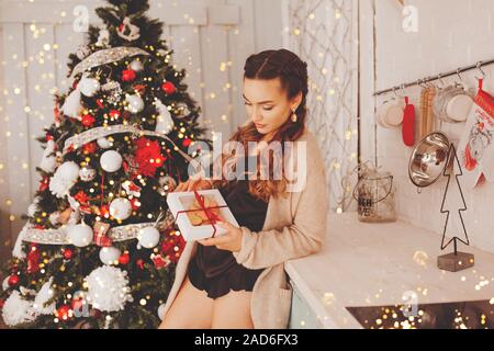 Young beautiful girl with pigtails in silk pajamas opens a box of cookies in the kitchen near the Christmas tree. Stock Photo