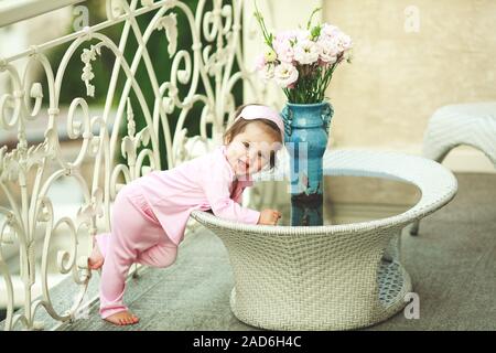 Cute little girl in pajamas, smiling stands near a table with flowers on a hotel balcony in summer Stock Photo