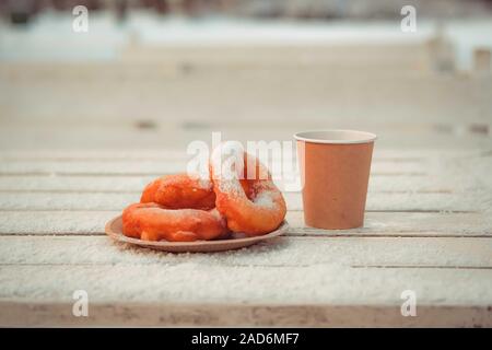 Russian fast food bagel pyshki and paper cup with tea or coffe on wooden table, covered with snow, winter snack in the park. Stock Photo