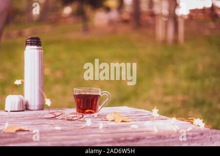 Cozy autumn tea break with a cup of tea , thermos flask,sunglasses, and dry autumn leaves on old vintage wooden table. top down view. Stock Photo