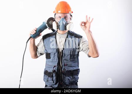 A builder in a helmet and a respirator, with an angle grinder on his shoulder. Shows okay sign. Stock Photo