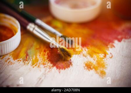 On a wooden art palette, smeared with bright red and yellow paints, lie two brushes and plastic lids from jars of paints. Stock Photo