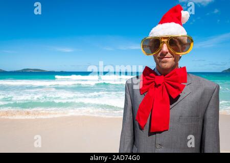 Portrait of a cheerful businessman in Santa hat, sunglasses and big red Christmas bow standing on a tropical beach Stock Photo