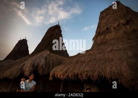 A young woman carrying a child as she is walking in front of traditional houses in traditional village of Tarung in West Sumba, Indonesia. Stock Photo