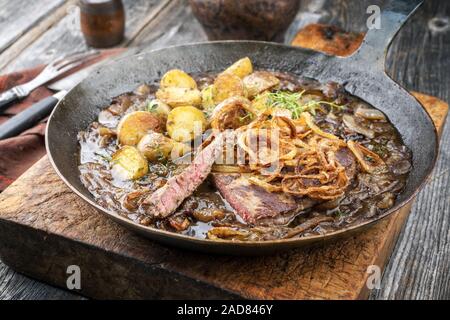 Traditional dry aged sliced roast beef with fried onion rings and potato chips as closeup in a wrough-iron pan with brown sauce Stock Photo