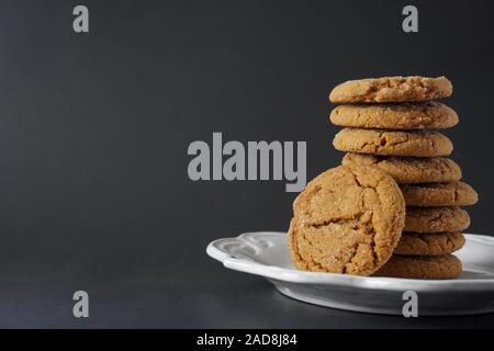 A gingerbread cookie leans against a stack of cookies on a white plate with a black background with copy space; landscape view Stock Photo