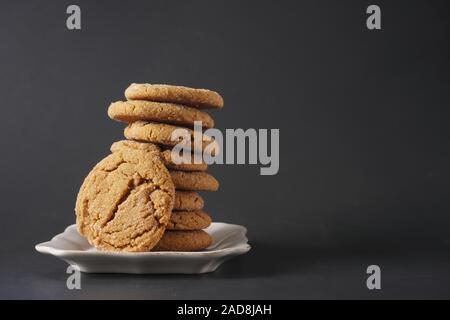 A crackly gingerbread cookie leans against a stack of cookies on a white plate with black background; copy space Stock Photo