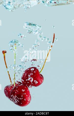 Close-up of two falling cherries followed by bubbles in front of bright blue background. Stock Photo