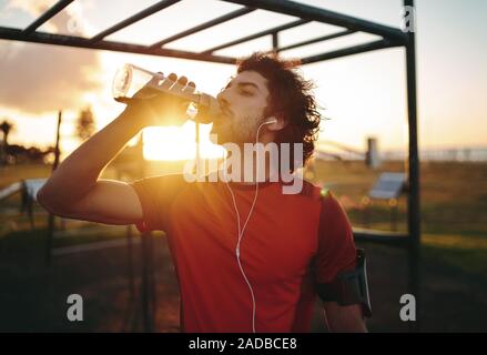 Fitness young athlete man with earphones in his ears drinking water from a reusable bottle after workout exercising on summer in gym park