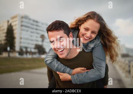 Cheerful young man carrying a woman on his back outdoors in the city street - laughing and having fun