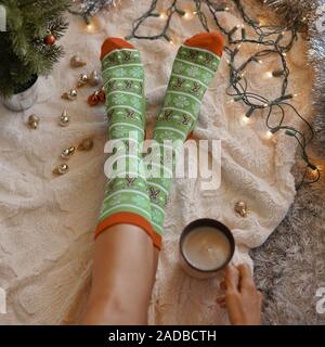 Feet in christmas socks near the Christmas tree. Woman sitting at the blanket, drinks hot beverage and relaxes warming up their feet in woollen socks. Stock Photo