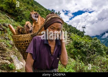 An old man is carrying a basket full with firewood, mountainous landscape in the distance Stock Photo