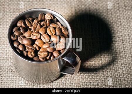 Metal mug full of roasted coffee beans on a hessian background. Stock Photo
