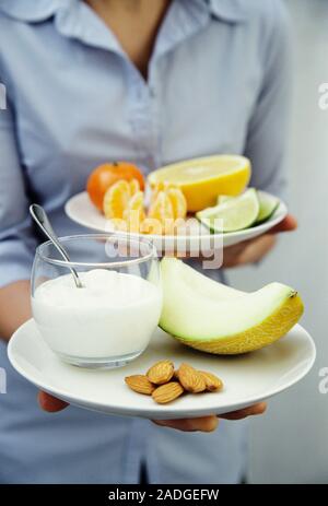 Healthy breakfast. Woman carrying a healthy breakfast. In her right hand she is holding a plate of yoghurt, a slice of melon (Cucumis melo) and shelle Stock Photo