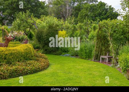 Sweeping path with herbaceous borders and seating area beneath a living Willow structure Stock Photo