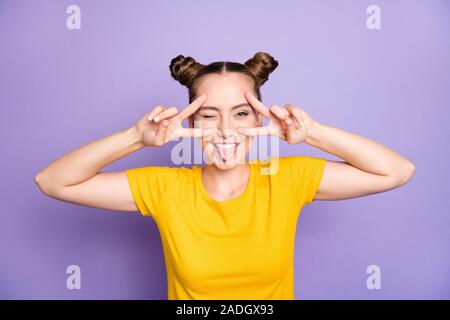 Photo of funky teenager lady holding hands near eyes showing v-sign symbols sticking tongue out mouth winking wear yellow t-shirt isolated on pastel Stock Photo