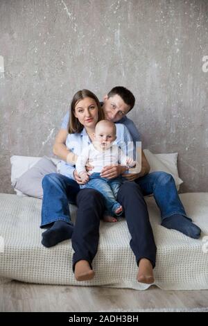 Happy Young Caucasian Family in Studio. Mother Holding Little Daughter in Arms. Father Hugging Her Wife and Baby Girl. People Sitting on Bed. Parents Stock Photo