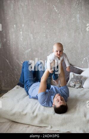 Happy Young Caucasian Family in Studio. Father is Holding and Hugging His Baby Daughter in Hands. Man Lying on Bed. Man Smiling. Infant Dissatisfied a Stock Photo