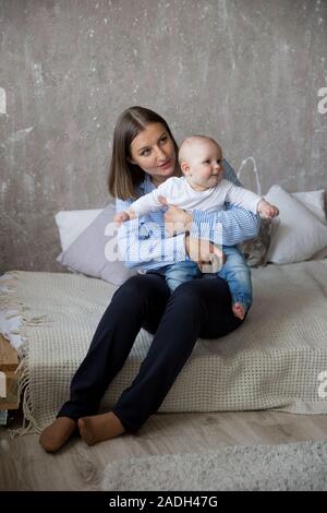 Happy Young Caucasian Family in Studio. Mother is Holding and Hugging His Baby Daughter in Hands. Beutiful People Sitting on Bed. Woman Smiling. Infan Stock Photo