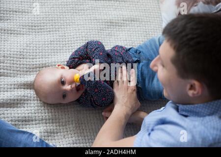 Happy Young Caucasian Family in Studio. Father is Touching His Baby Daughter. People Lying on Bed. Man Smiling. Infant Serious. Infant Holding Toy Ban Stock Photo