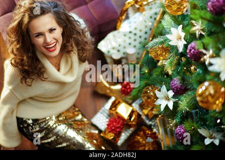 Upper view of smiling trendy 40 year old housewife with long brunette hair in gold sequin skirt and white sweater under decorated Christmas tree near Stock Photo