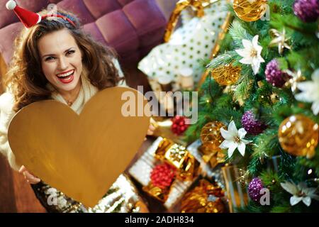 Upper view of smiling trendy woman with long brunette hair in gold sequin skirt and white sweater under decorated Christmas tree near present boxes si Stock Photo