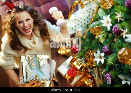 Smiling trendy housewife with long brunette hair in gold sequin skirt and white sweater under decorated Christmas tree near present boxes holding torn Stock Photo