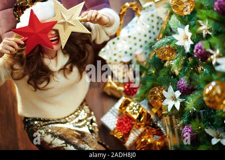 Cheerful trendy housewife with long brunette hair in gold sequin skirt and white sweater under decorated Christmas tree near present boxes holding 2 r Stock Photo