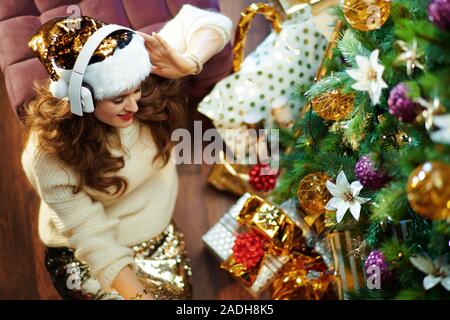 Upper view of smiling trendy woman with long brunette hair in gold sequin skirt and white sweater listening to the music with headphones under decorat Stock Photo