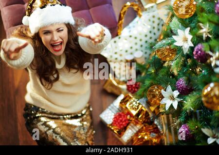 Upper view of smiling modern 40 year old woman with long brunette hair in gold sequin skirt and white sweater under decorated Christmas tree near pres Stock Photo